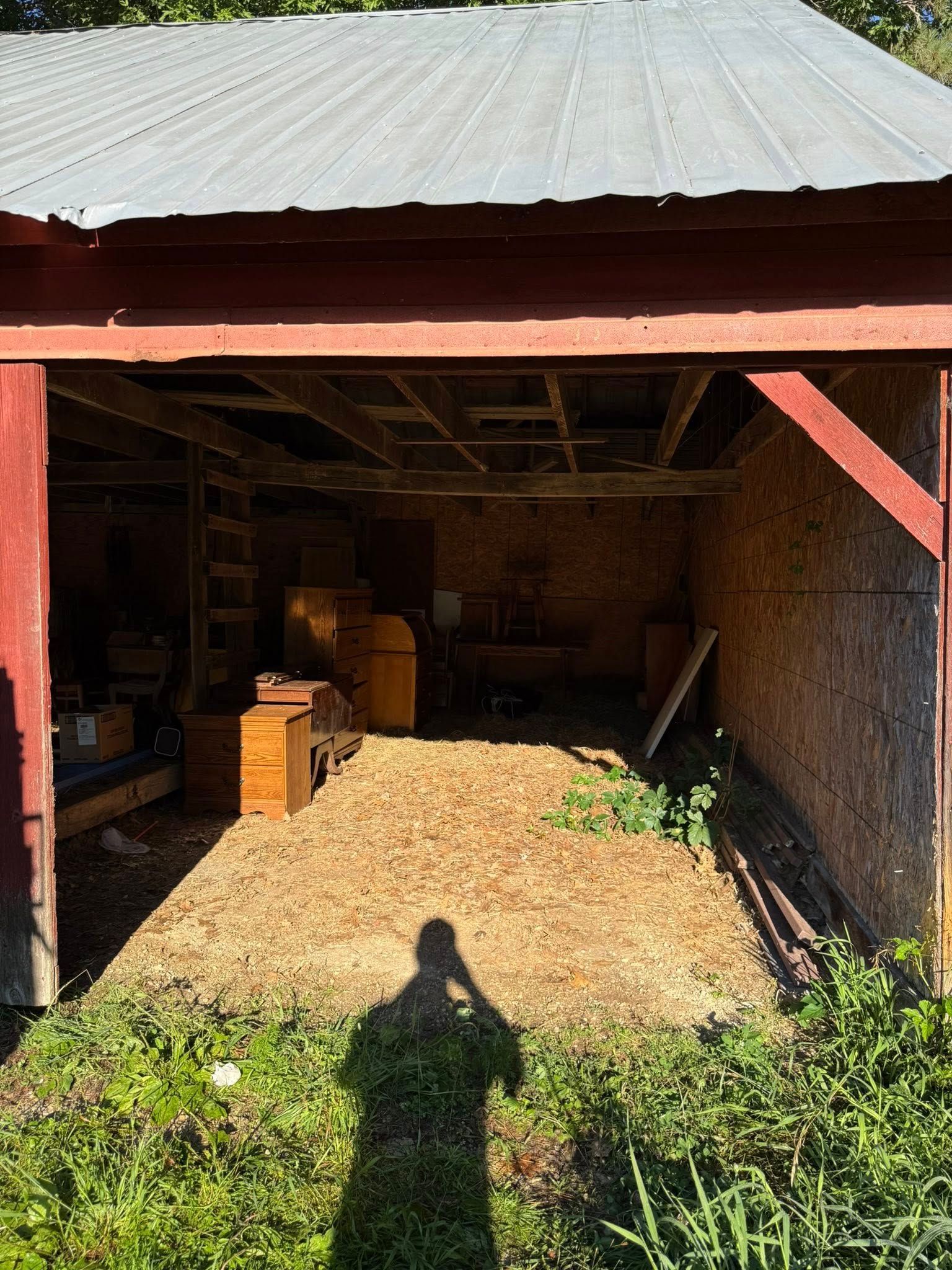 An open, red-framed shed with a metal roof, showing a dirt interior with stored wooden items and tools.