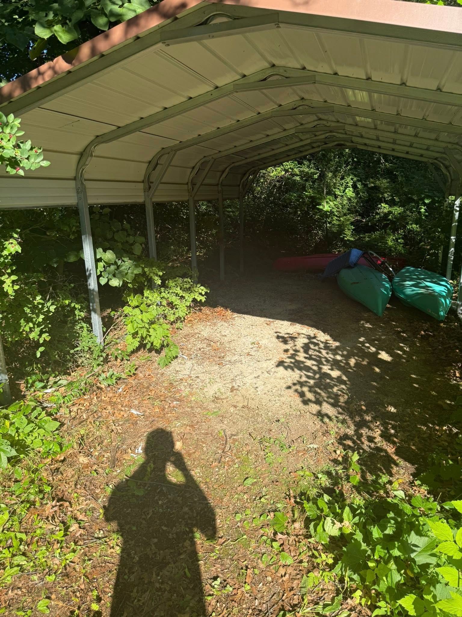 A metal carport stands in a wooded area with several colorful canoes and kayaks resting underneath on gravel.