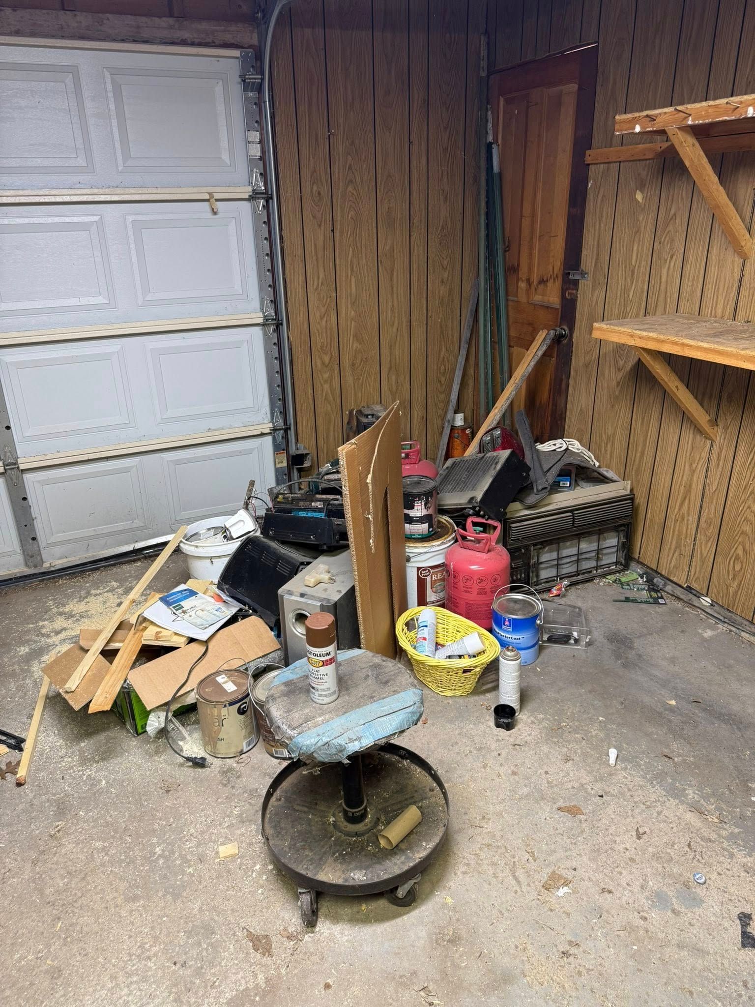 A cluttered garage interior with a rolling stool, cardboard boxes, various tools, and a wooden shelf on a pegboard wall.