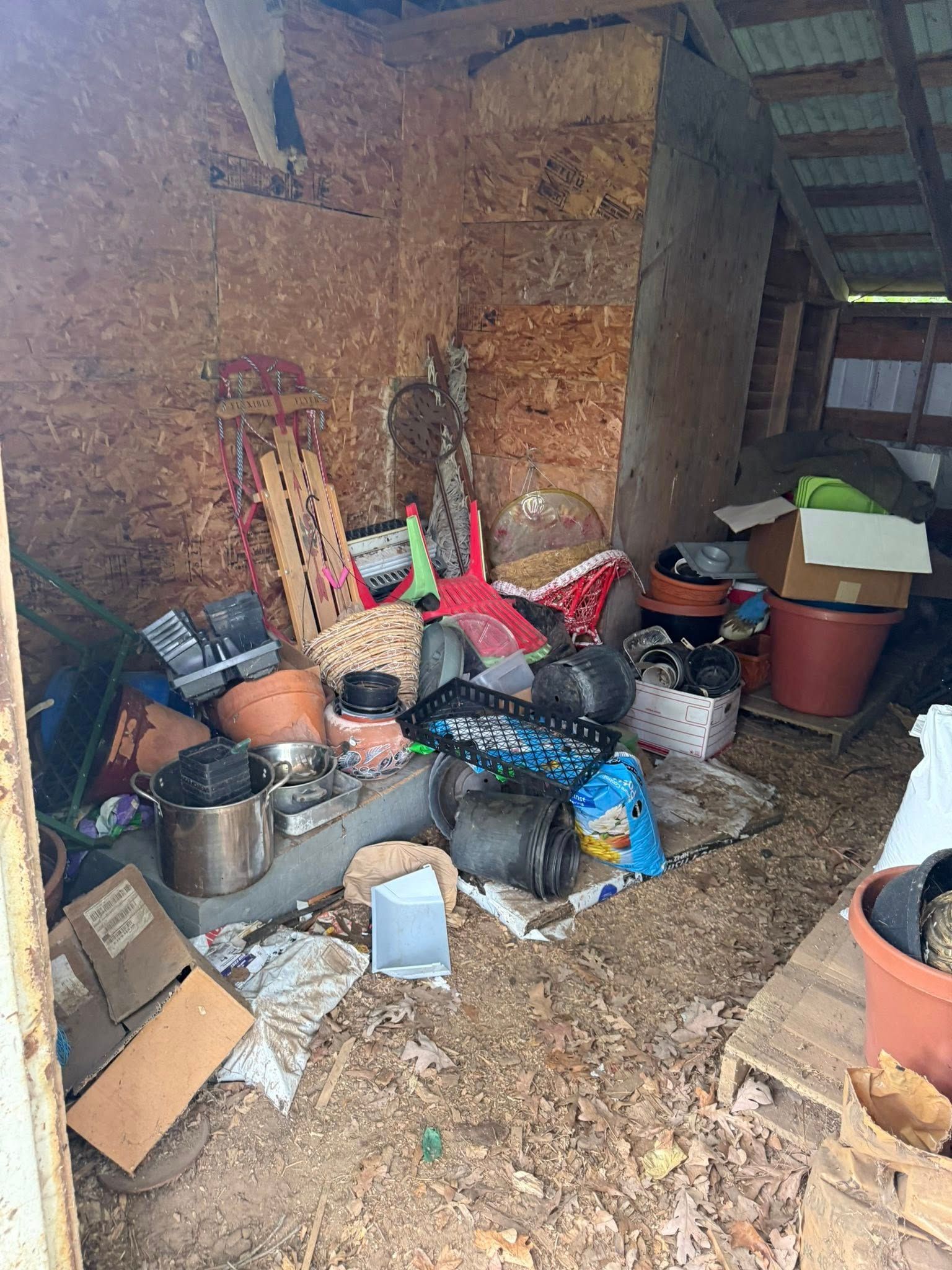 A cluttered shed interior filled with various plastic pots, garden tools, and cardboard boxes on a dirt floor.