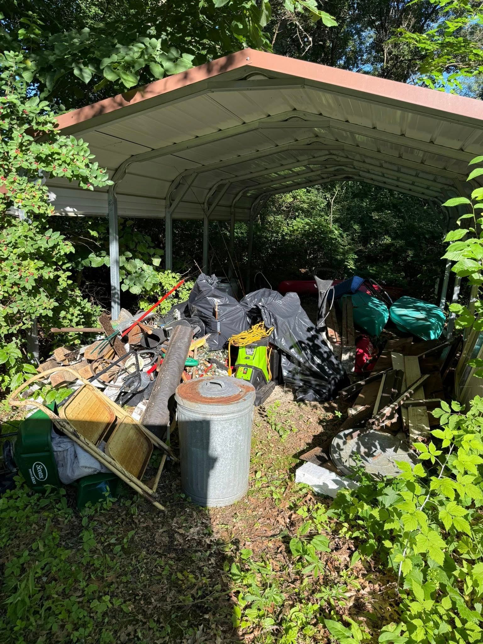A metal carport filled with miscellaneous items, including tools, a plastic bucket, and discarded household clutter.