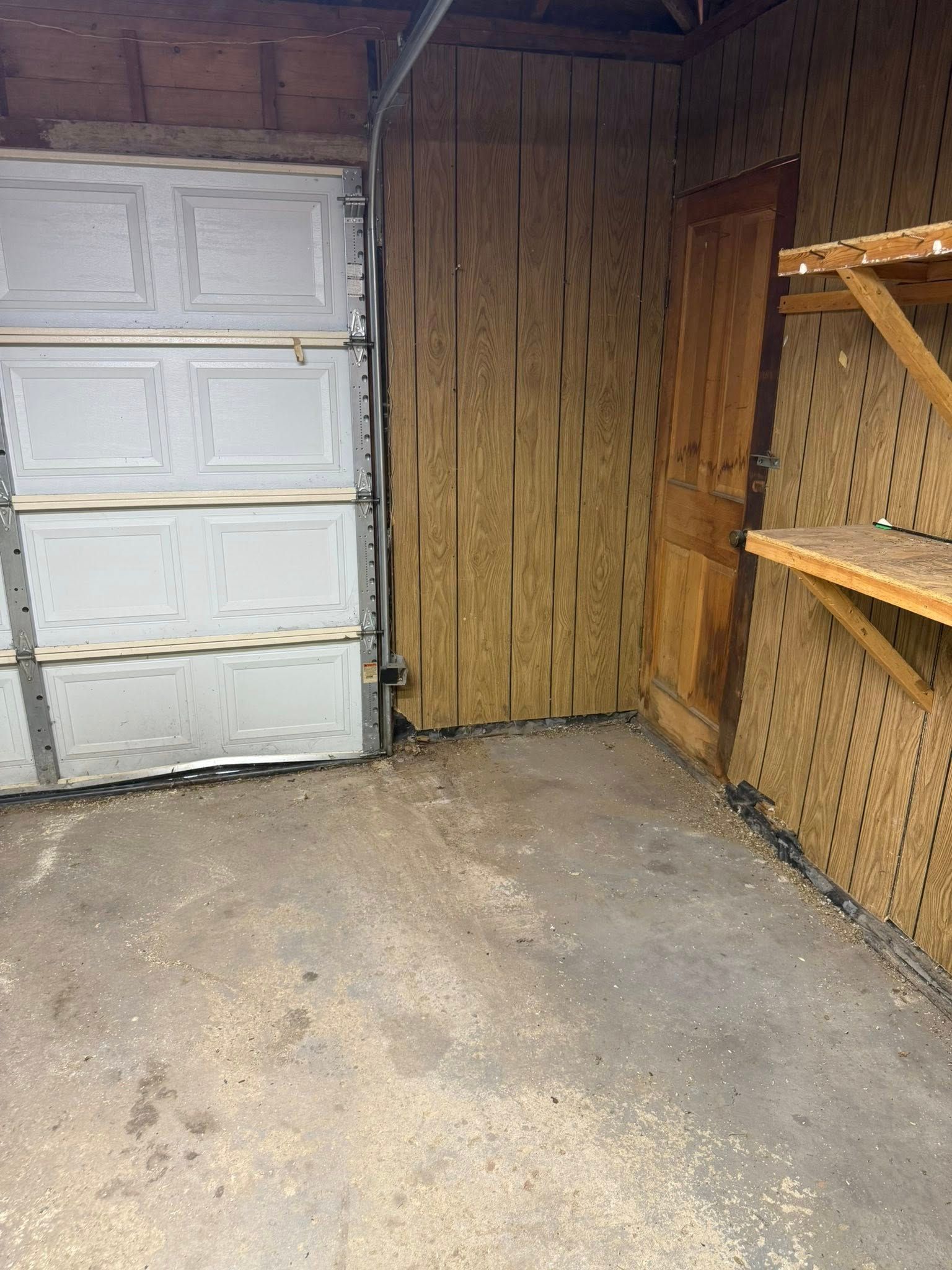 A garage interior with a white sectional door, plywood paneled walls, a wooden door, and a wall-mounted shelf.