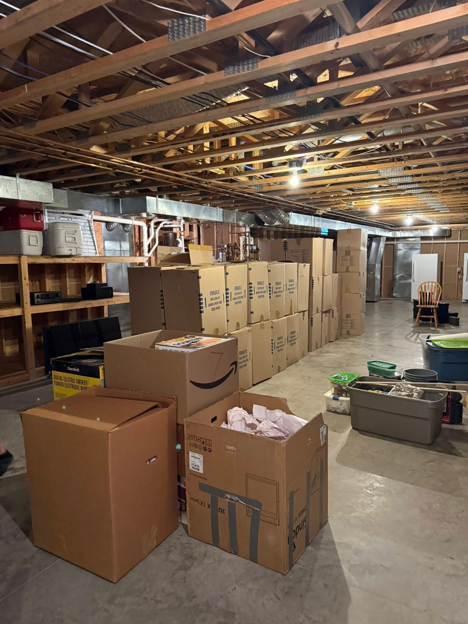 Unfinished basement filled with cardboard moving boxes stacked on a concrete floor near wooden shelving and exposed beams.