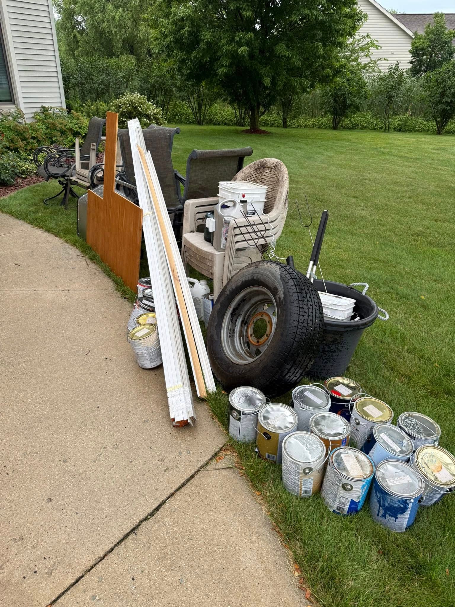 A pile of junk including furniture, a tire, and multiple paint cans sitting on a lawn next to a concrete sidewalk.