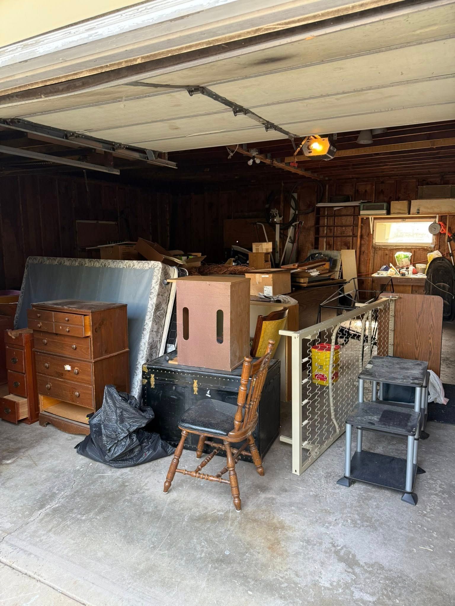A garage interior filled with various items, including a mattress, dresser, cardboard box, wooden chair, and storage bins.