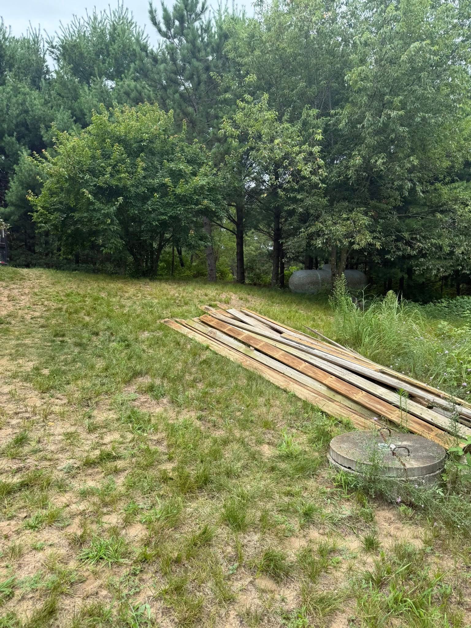 A stack of wooden planks lies on a patch of grass near a concrete septic tank cover, with trees in the background.