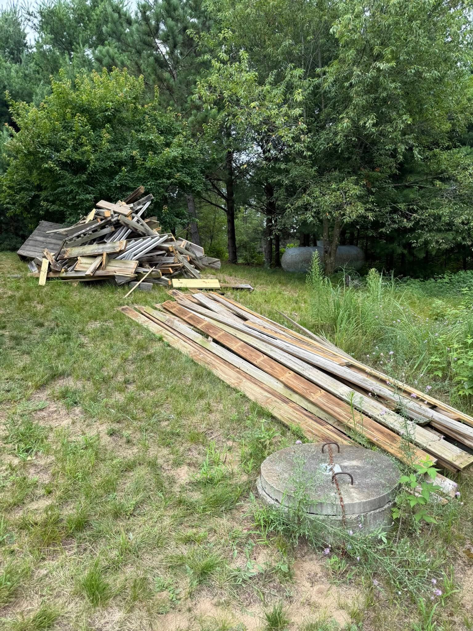 A pile of construction debris and a stack of long wooden boards lie on a grassy field near trees.