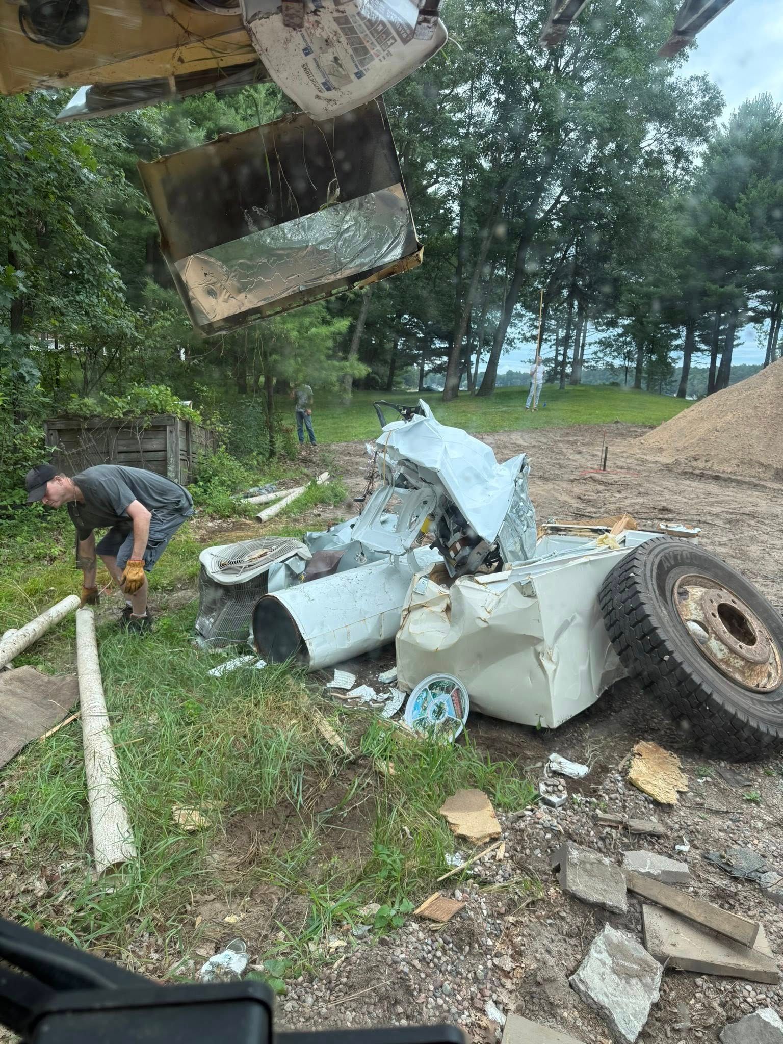A person stands near a pile of scrap metal and a large tire outdoors, with a mechanical arm hovering overhead.