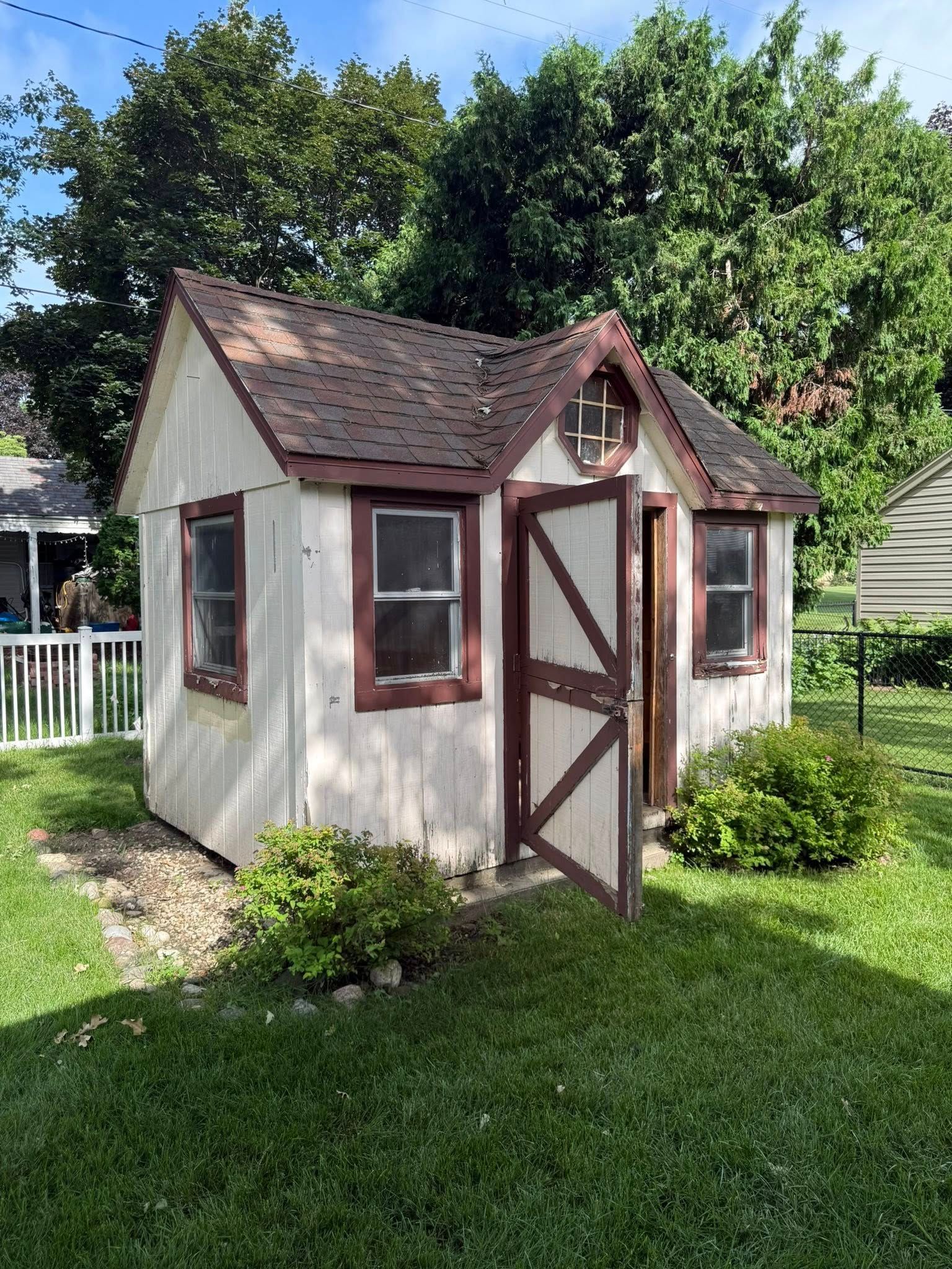 A small, white wooden shed with dark red trim, a gabled roof, and a partially open door, situated in a green backyard.