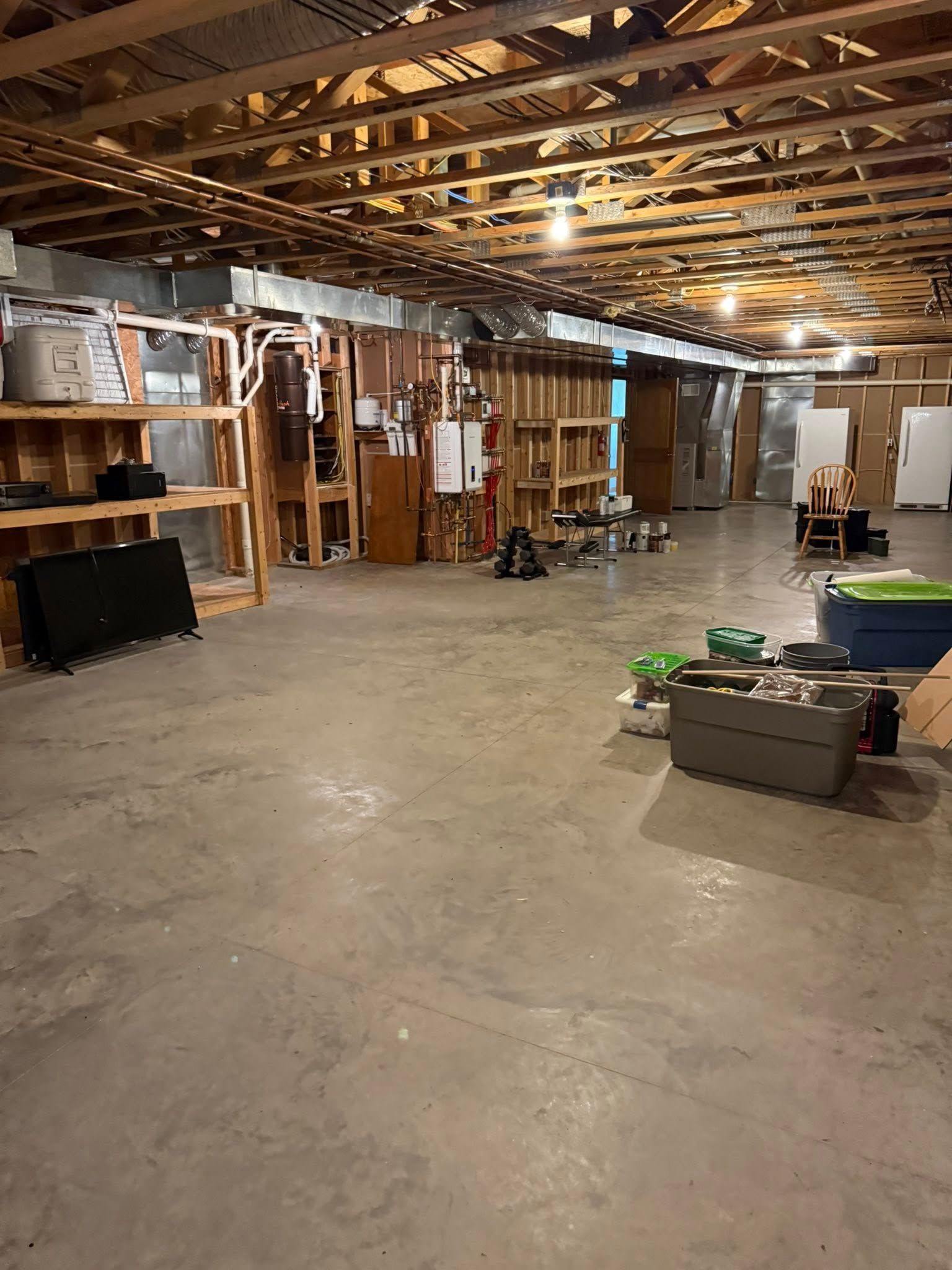 An unfinished basement with exposed wooden ceiling joists, concrete floors, wooden shelving, and stored items.