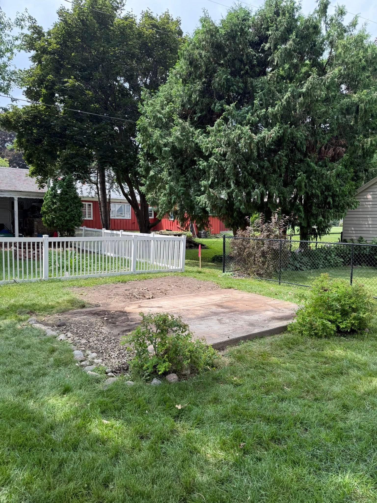 A rectangular patch of dirt and gravel sits in a grassy backyard, surrounded by trees, a white fence, and a red house.
