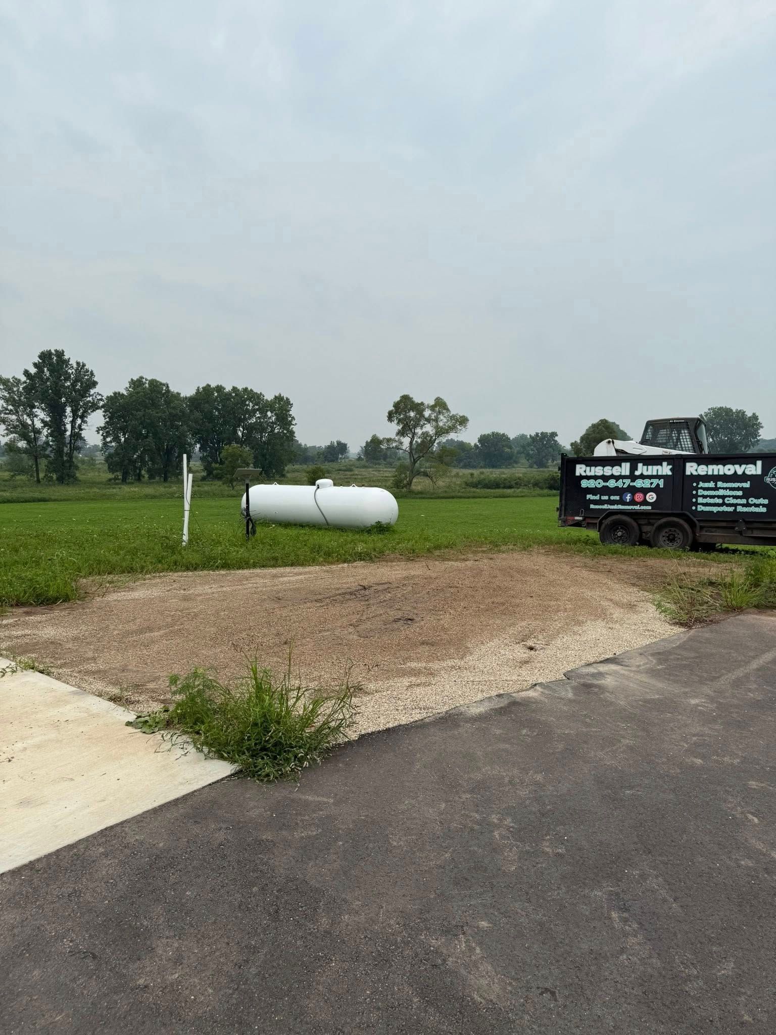 A white propane tank sits in a grassy field next to a utility truck parked on a gravel patch near an asphalt road.