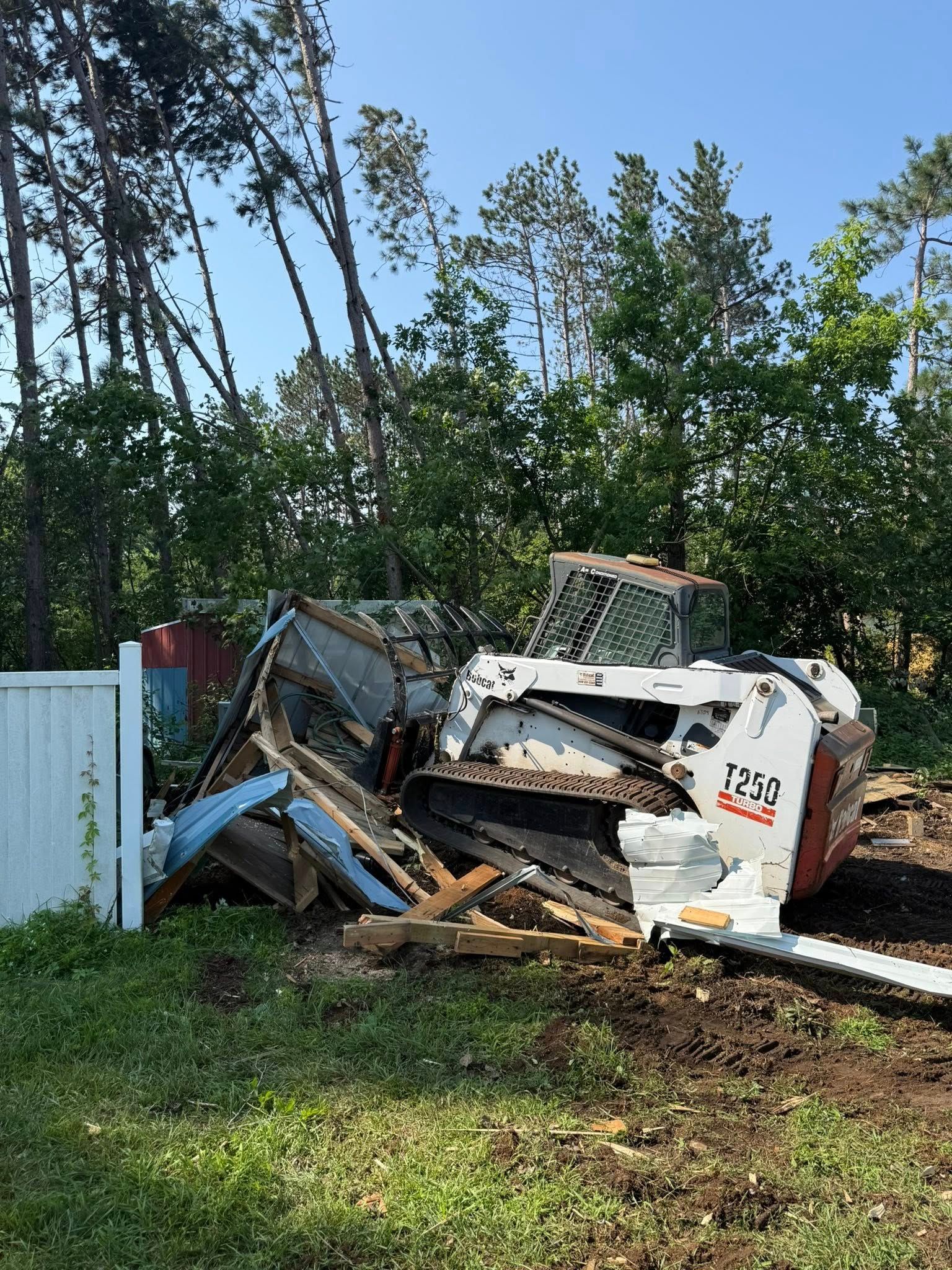 A white Bobcat compact track loader sits amidst a pile of debris and twisted metal near a white fence and line of trees.