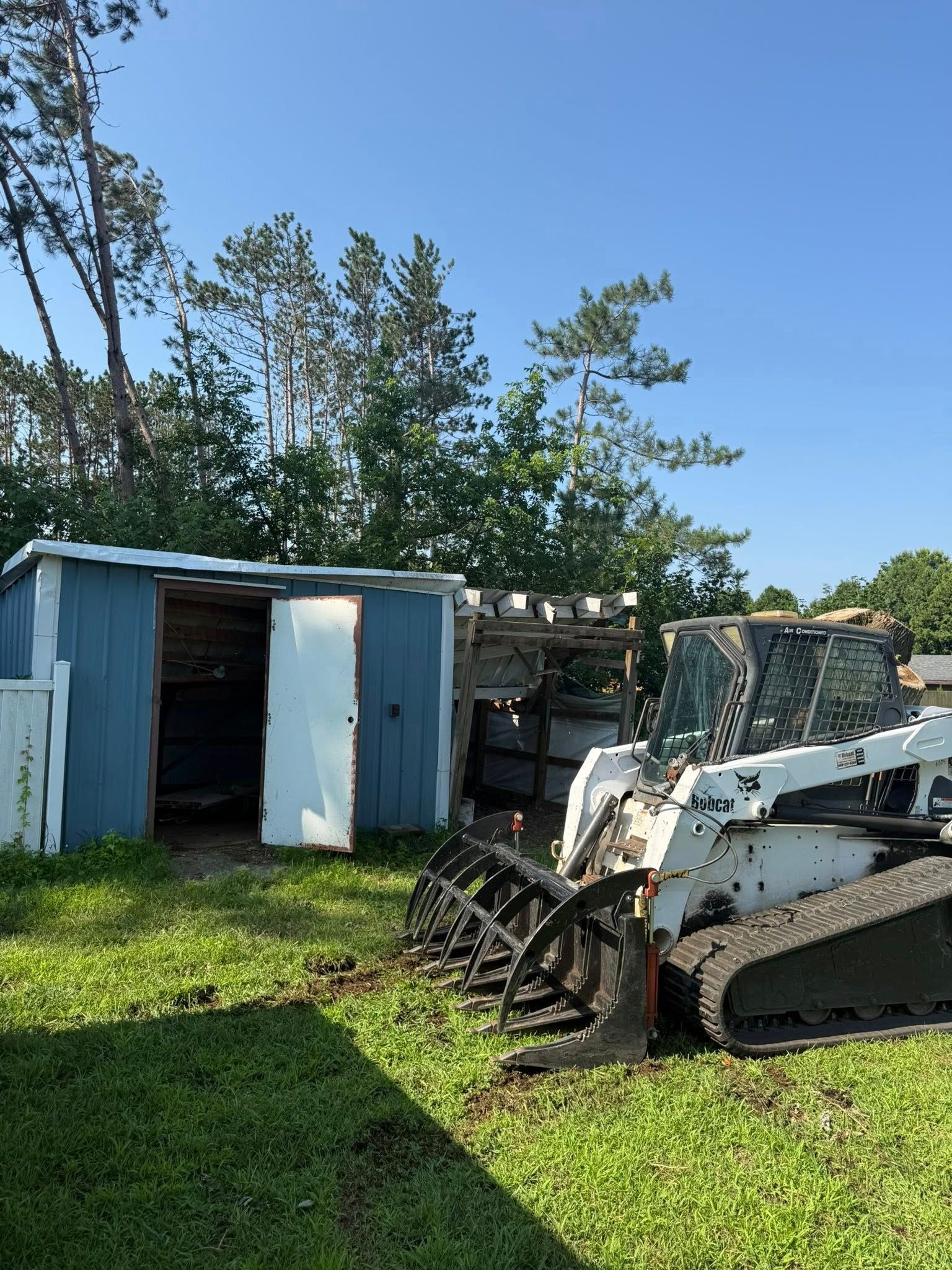 A white tracked skid steer with a grapple attachment parked on a grassy lawn next to a small blue storage shed.