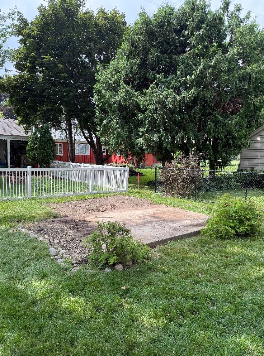A concrete pad sits in a grassy backyard, surrounded by trees and a white fence, with a red building in the background.