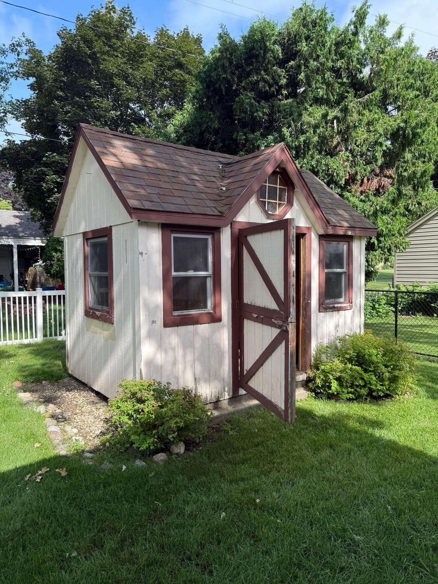A small, white, cottage-style wooden shed with brown trim and an open door, set in a backyard with grass and trees.