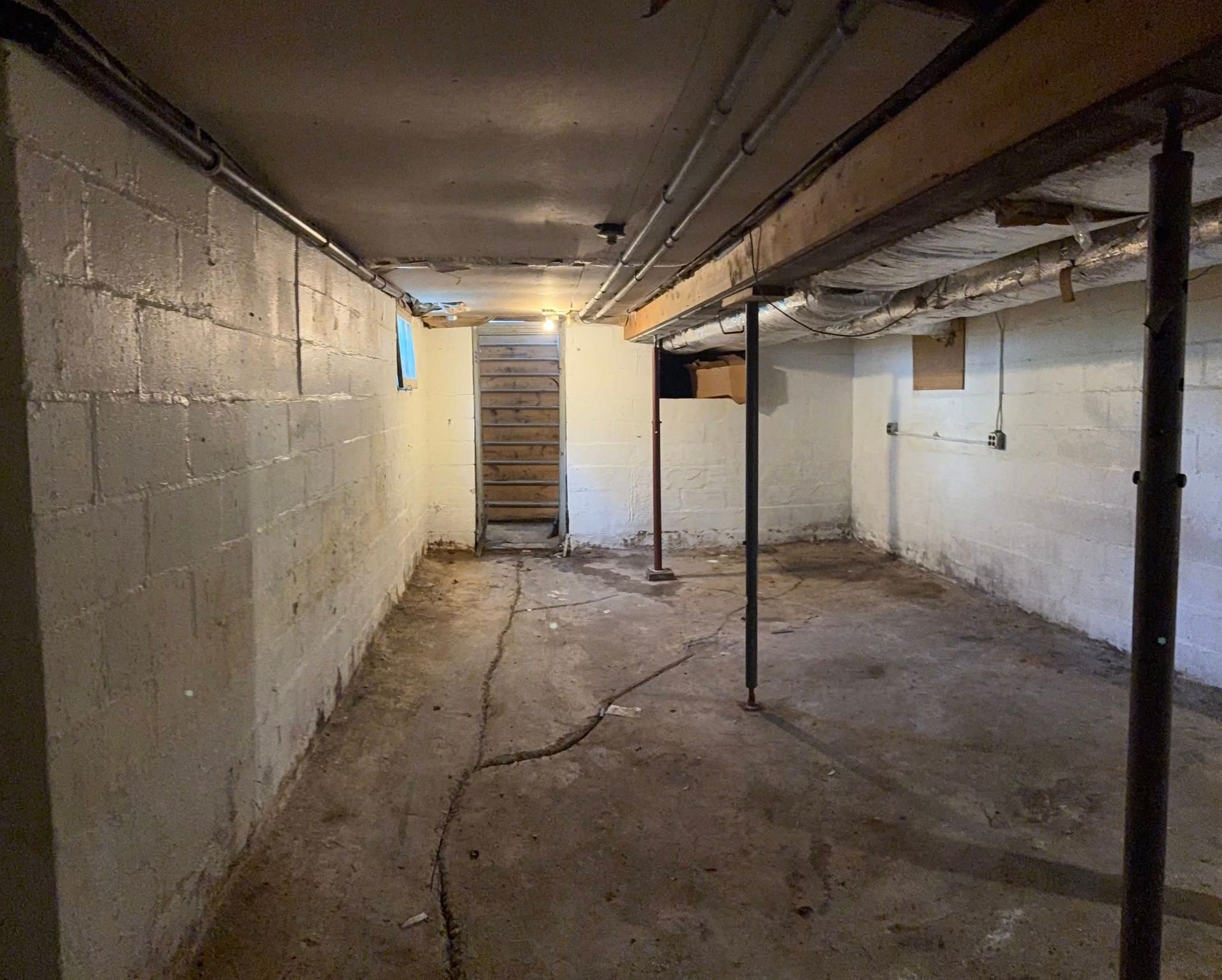 An unfinished basement with white concrete block walls, a concrete floor with cracks, and metal support posts.
