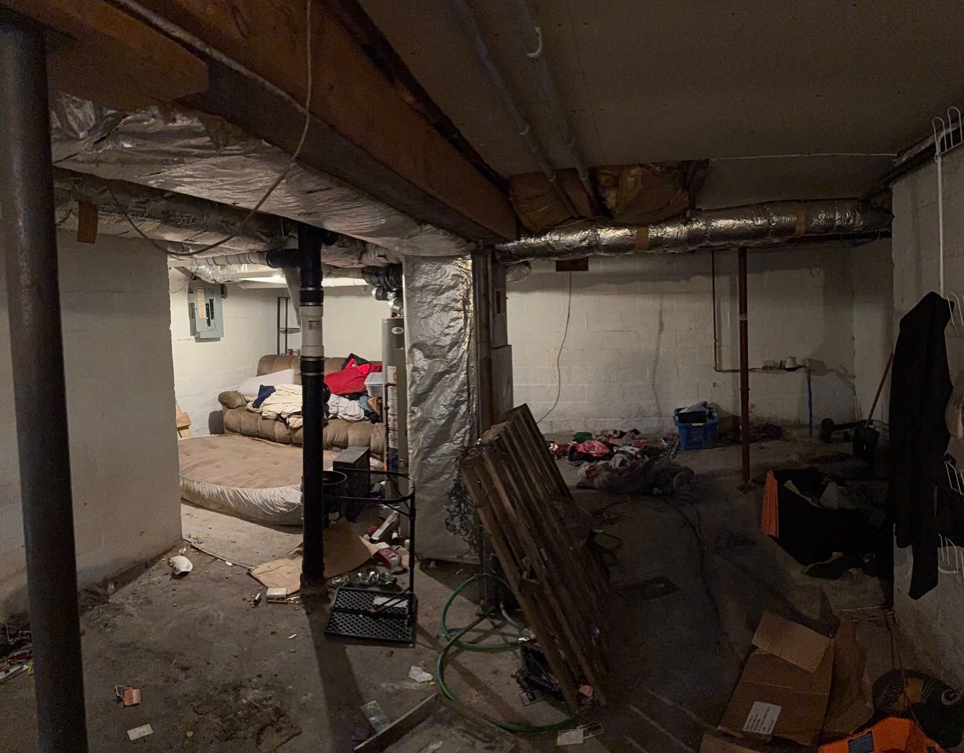 A dimly lit, unfinished basement with concrete floors, exposed ceiling beams, a couch in the back, and scattered items.