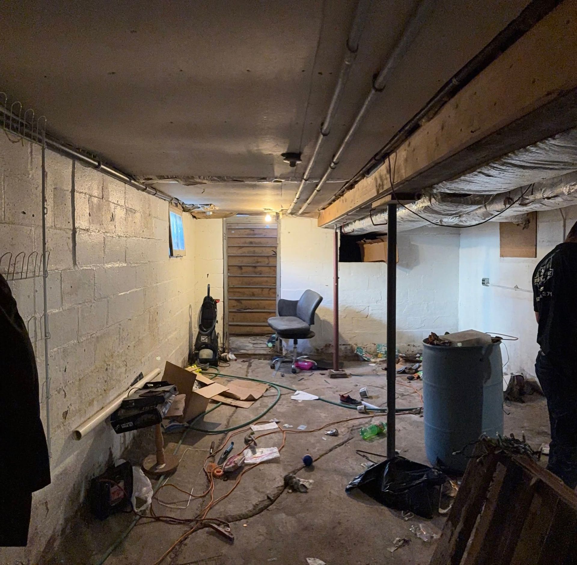 A cluttered, unfinished basement with concrete block walls, a metal support pole, a desk chair, and scattered debris.