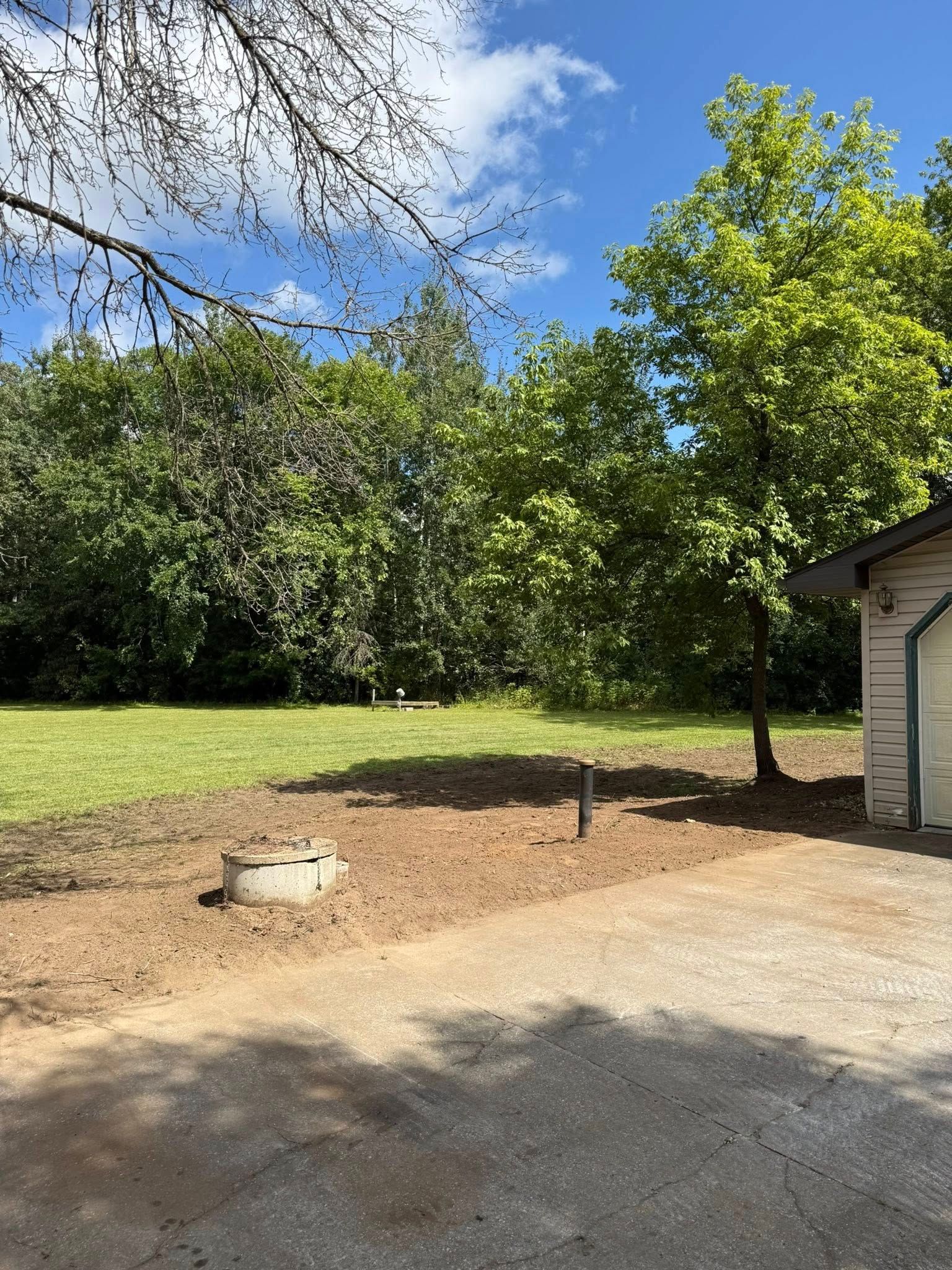 A sunlit yard with a circular concrete structure, bare soil in the foreground, grassy field, and trees against a blue sky.