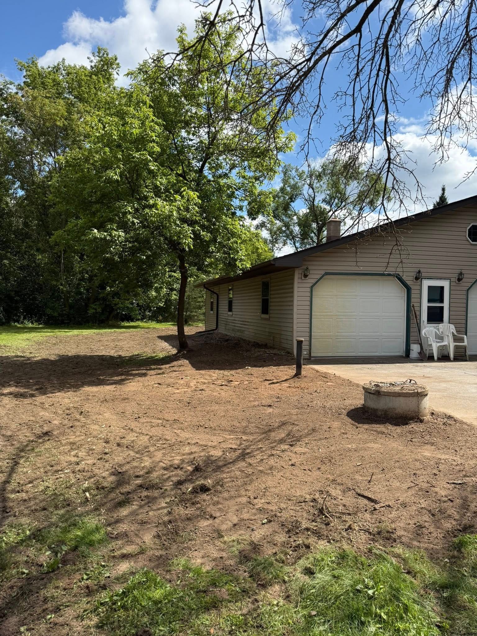 A beige house with a white garage door stands next to a large tree and a patch of cleared, dry ground under a sunny sky.