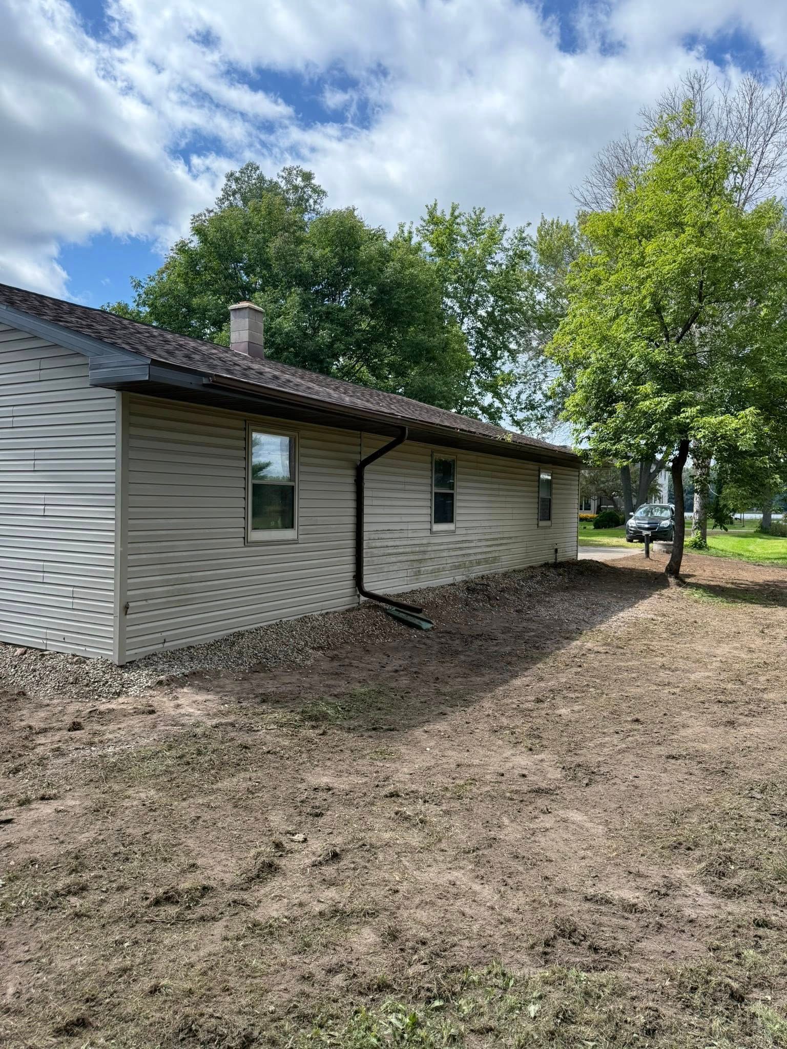 Light-colored, horizontal siding house exterior with a dark roof and downspout, bordered by a dirt yard and trees.