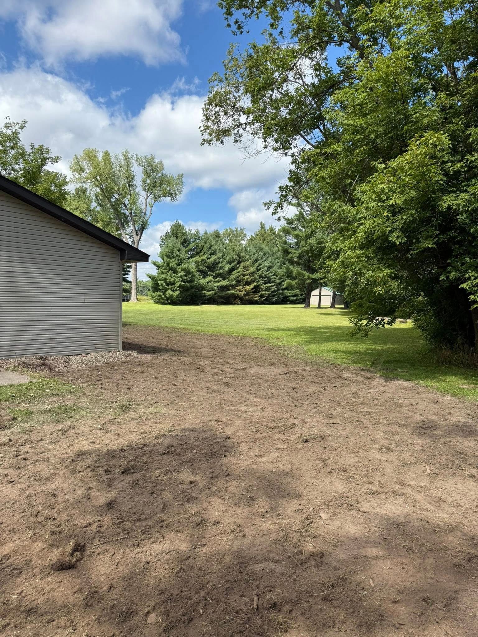 An expanse of dark, tilled soil borders a grey house siding, leading toward a grassy yard, trees, and a blue sky.