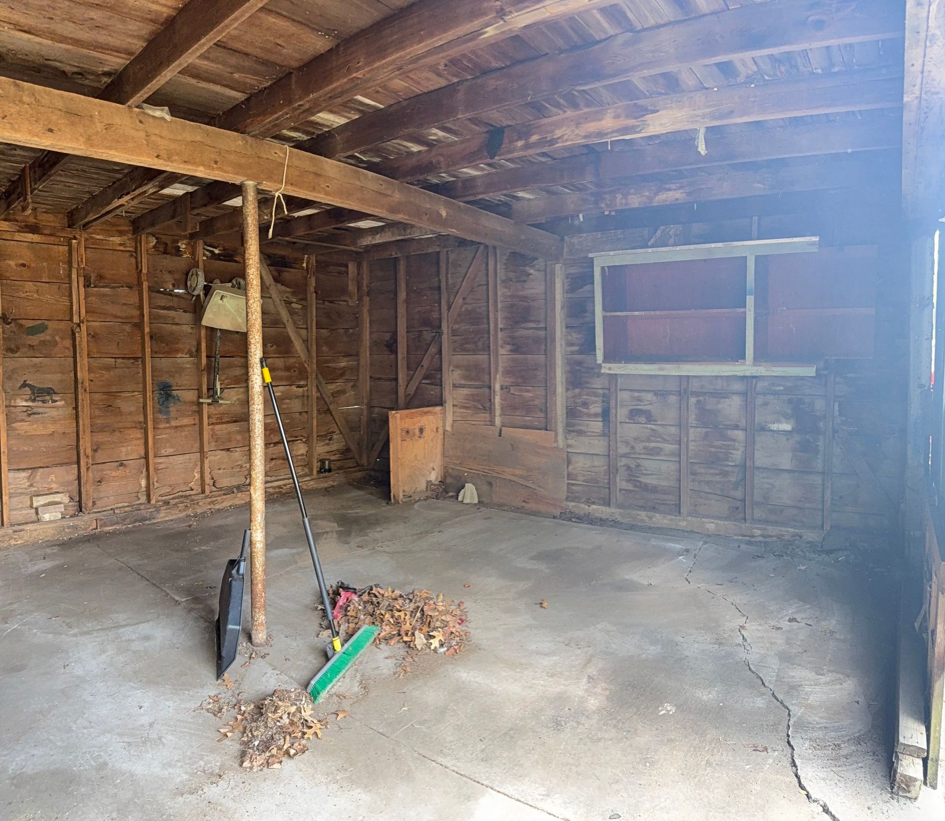 Interior view of a rustic, unfinished garage with wood walls, a concrete floor, exposed ceiling beams, and a window.