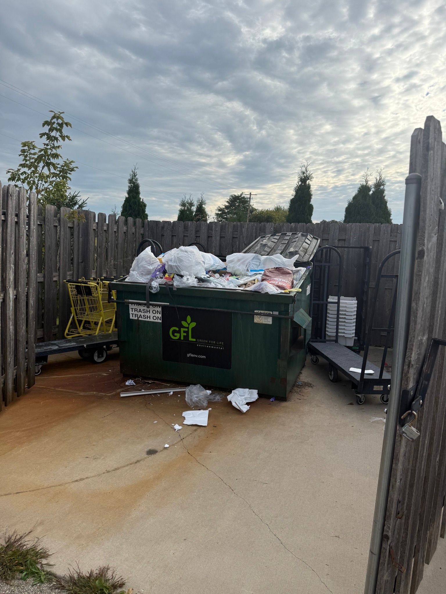A green dumpster filled with white trash sits in an outdoor enclosure with a wooden fence under a cloudy sky.