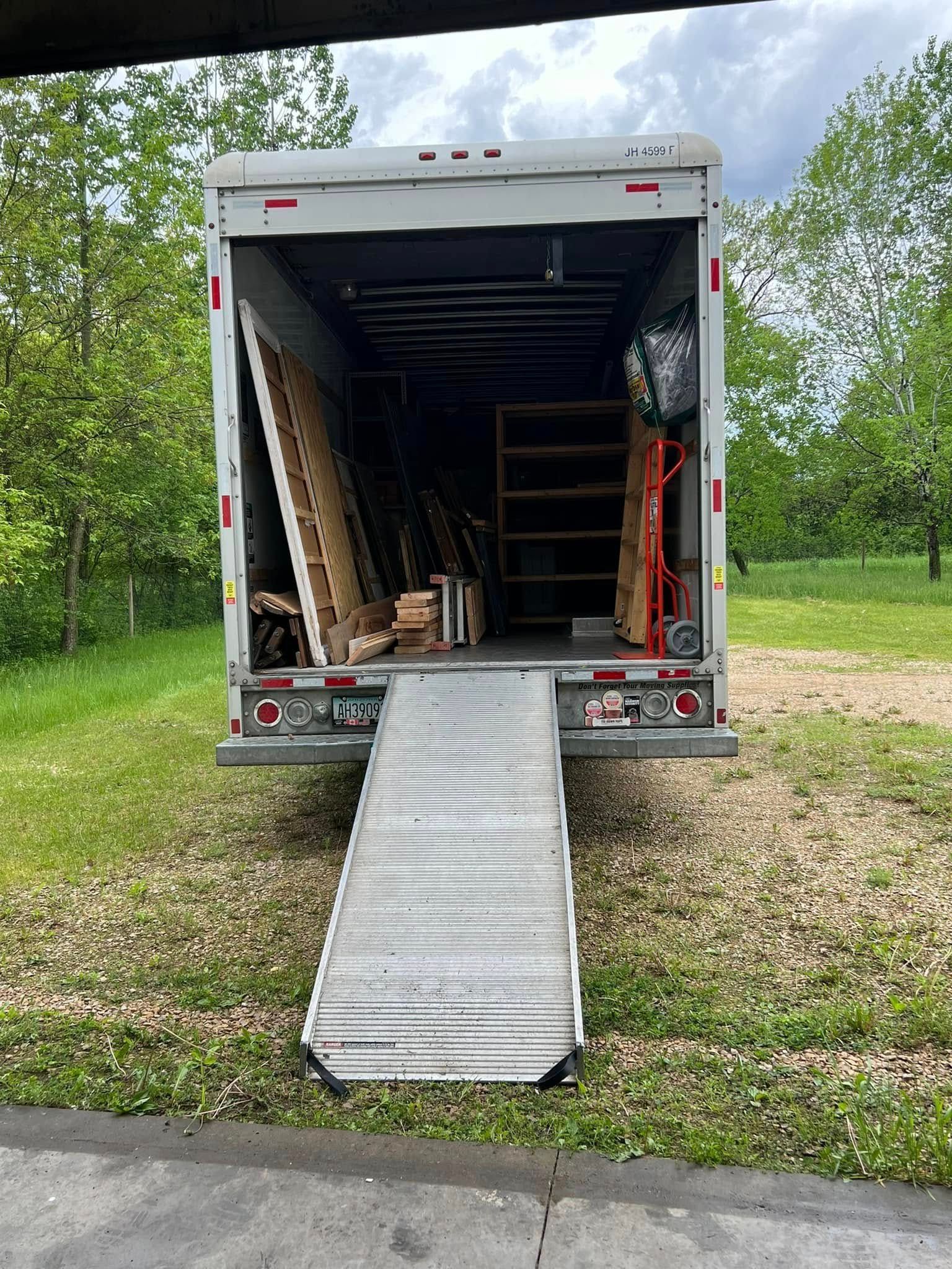 An open box truck parked on a gravel lot with a loading ramp extended to the ground, showing shelves and wood inside.