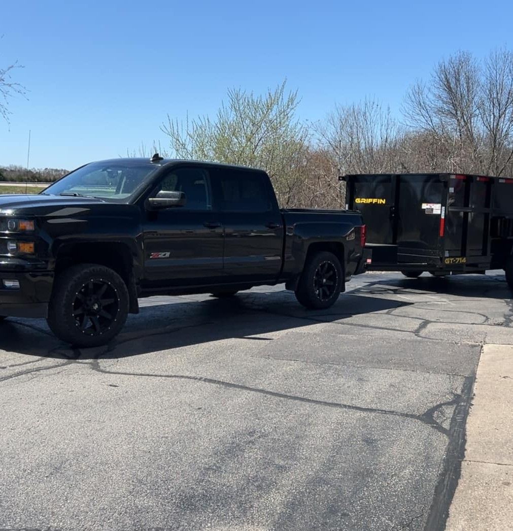 A black pickup truck parked on an asphalt lot, towing a black utility dump trailer on a clear, sunny day.