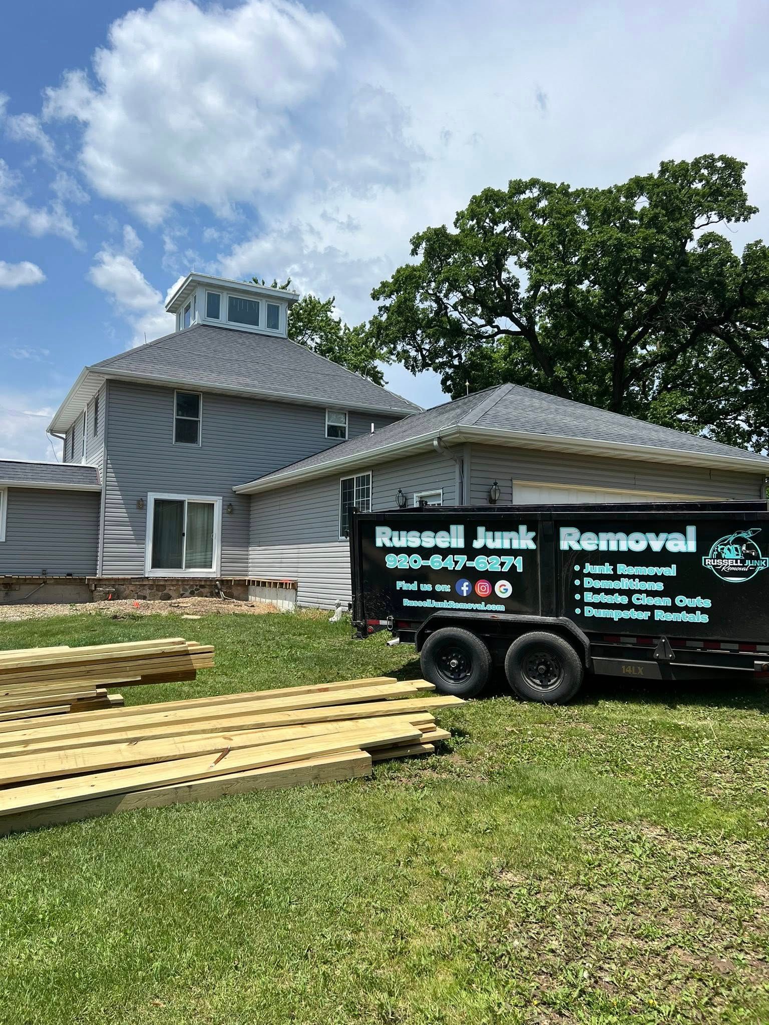 A Russell Junk Removal trailer parked on a lawn in front of a house undergoing exterior renovation.