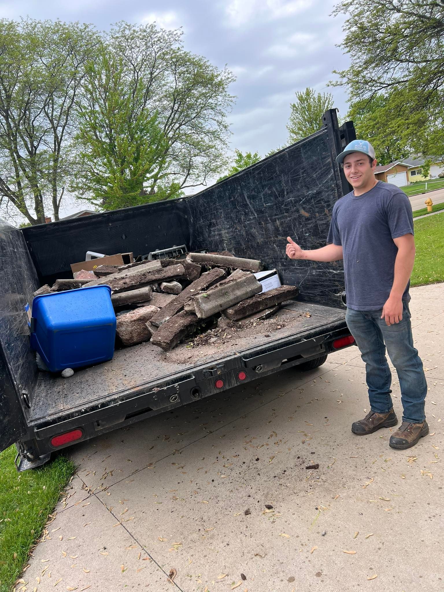 A person in a blue shirt and cap gives a thumbs up next to a dumpster filled with debris and a blue plastic bin.