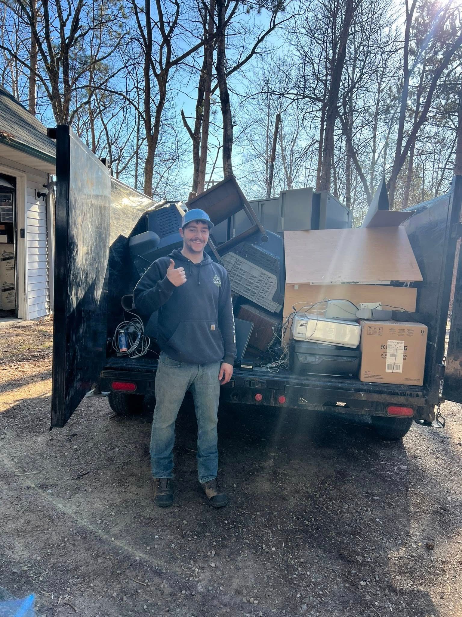 A person in a hoodie and cap gives a thumbs up in front of a dump trailer filled with household items and boxes.