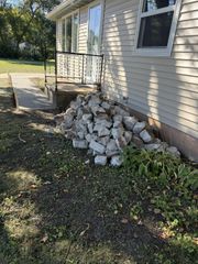 A pile of broken concrete chunks rests against the siding of a house, next to a porch and walkway on a sunny day.