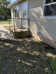 Tan vinyl-sided house with a small concrete porch, black metal railing, and a door surrounded by dead leaves and debris.