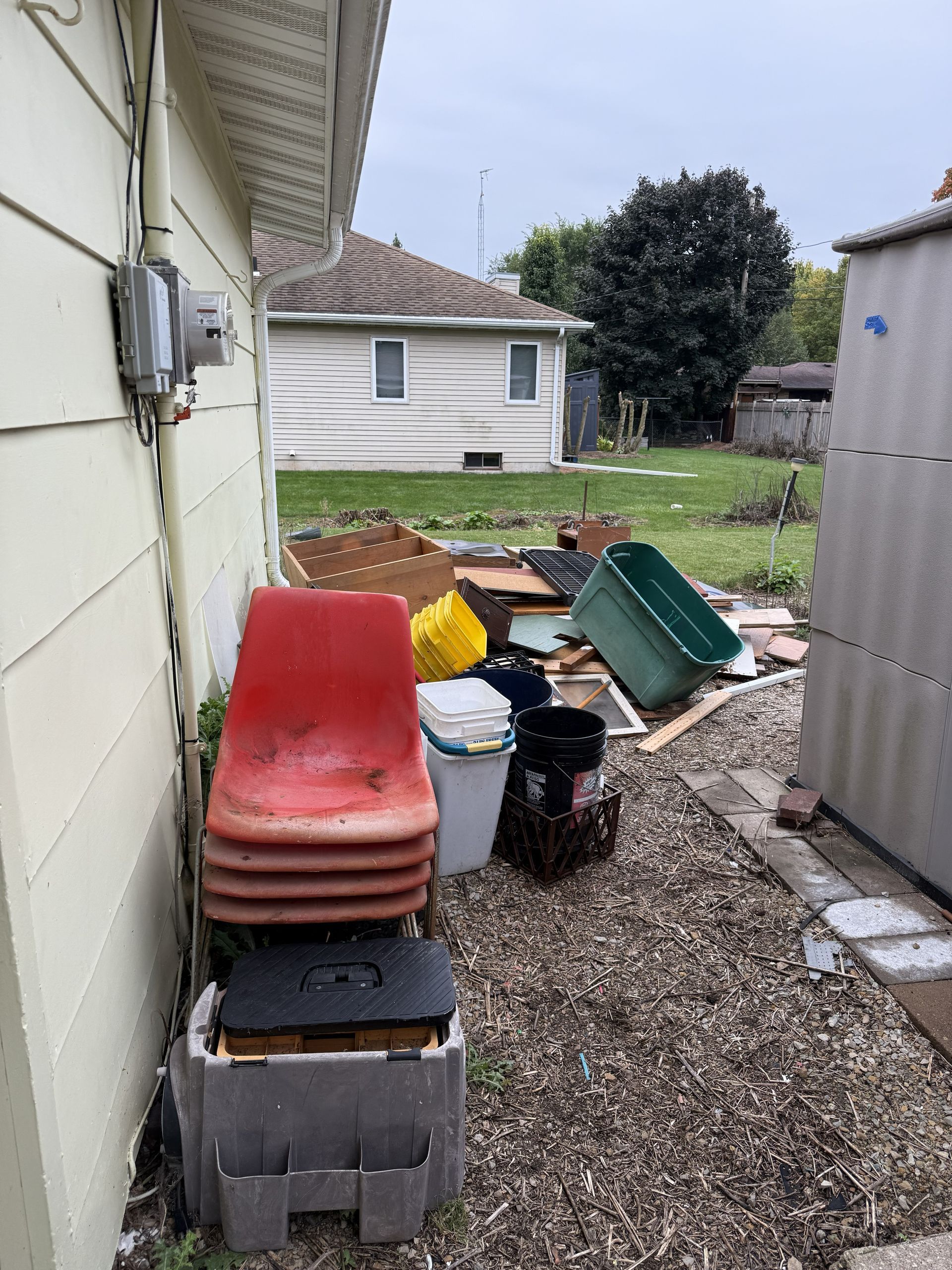 A collection of cluttered items, including red chairs, plastic bins, and cardboard, sits in a gravel side yard.
