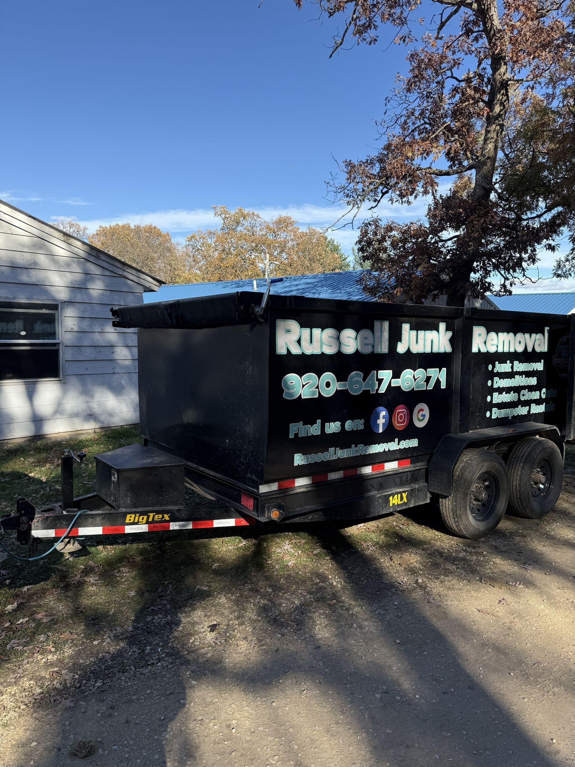 A black junk removal trailer parked on a gravel driveway next to a white building under a sunny sky.