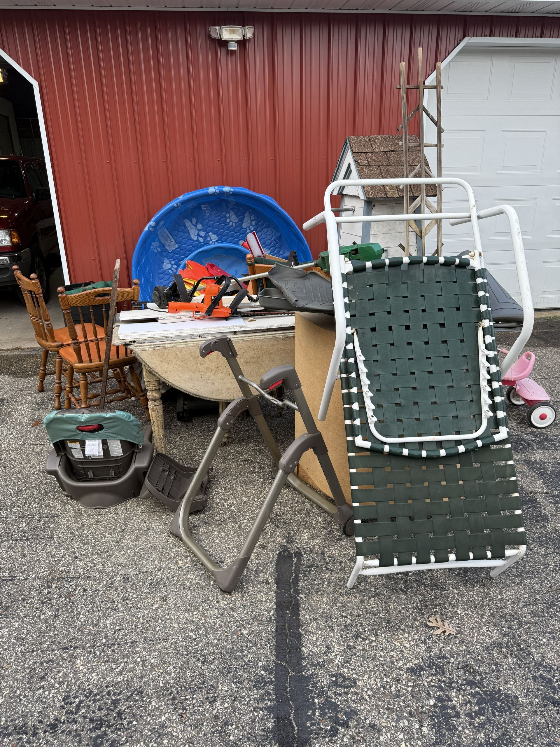 Assorted items, including a folding chair, plastic pool, and wooden chair, are gathered in front of a red building.