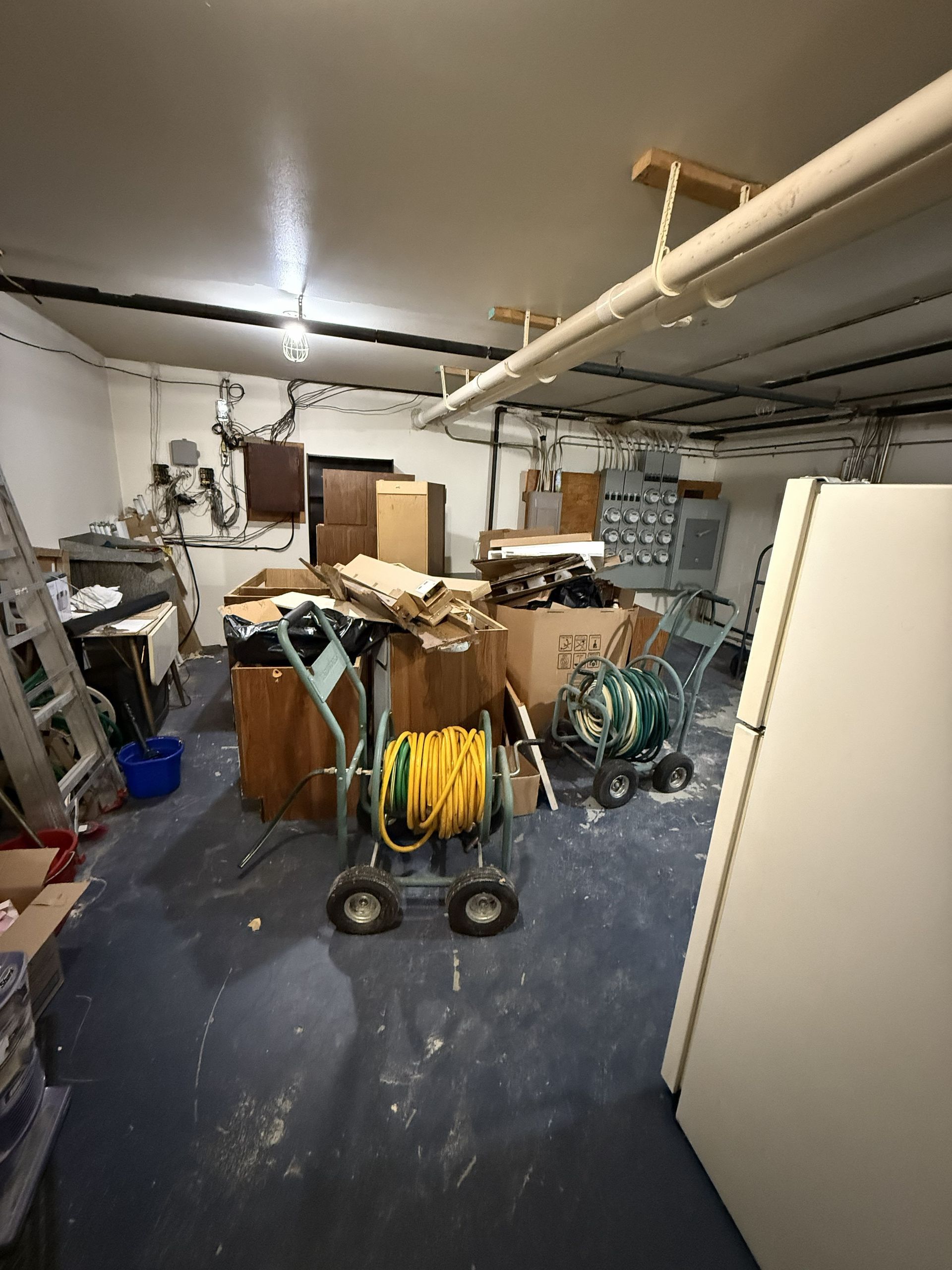 A cluttered garage featuring a yellow garden hose on a cart, cardboard boxes, various tools, and a large white refrigerator.