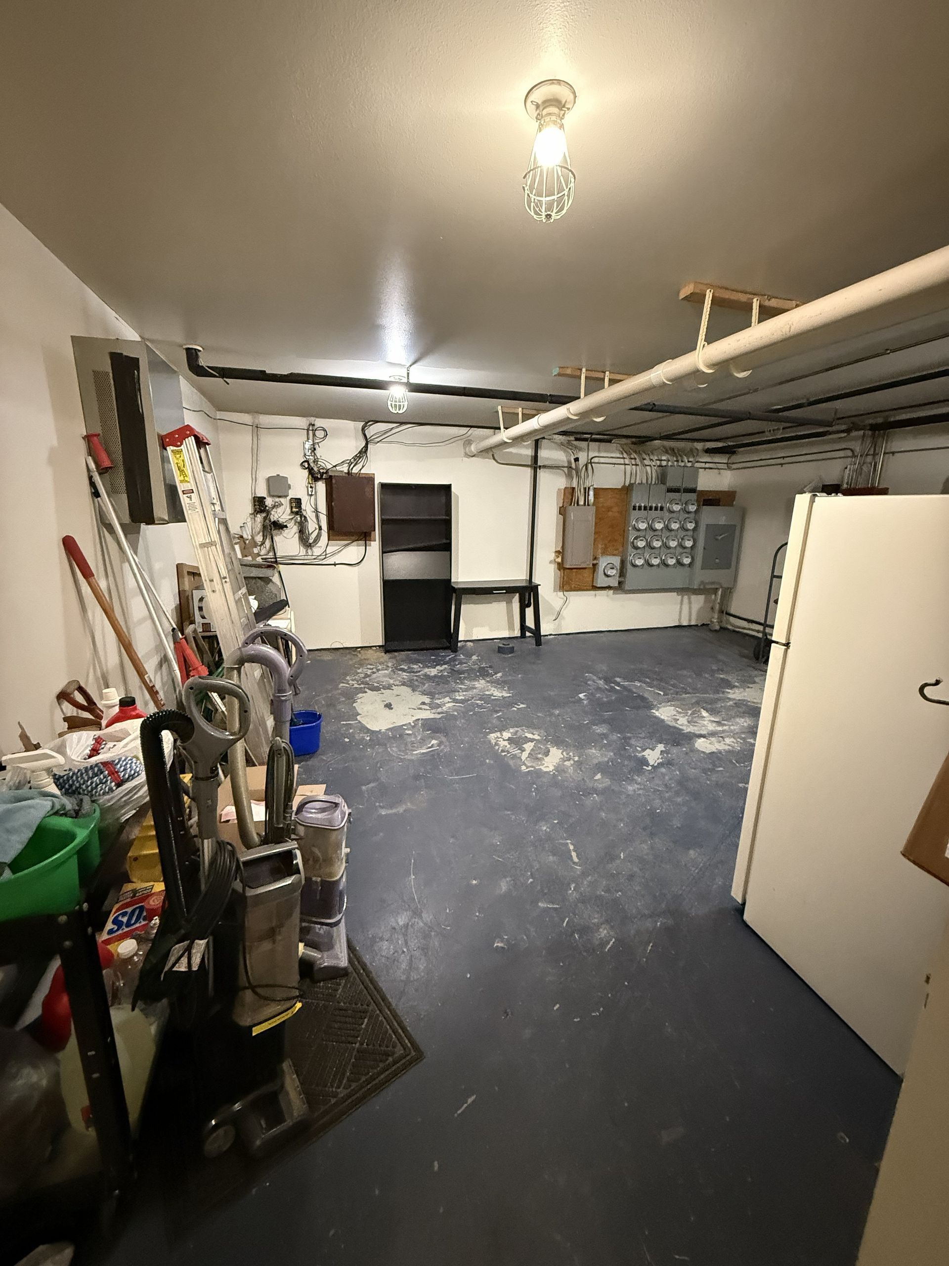A basement room features a gray floor with white paint splatters, cleaning tools, a shelf, and electrical panels.