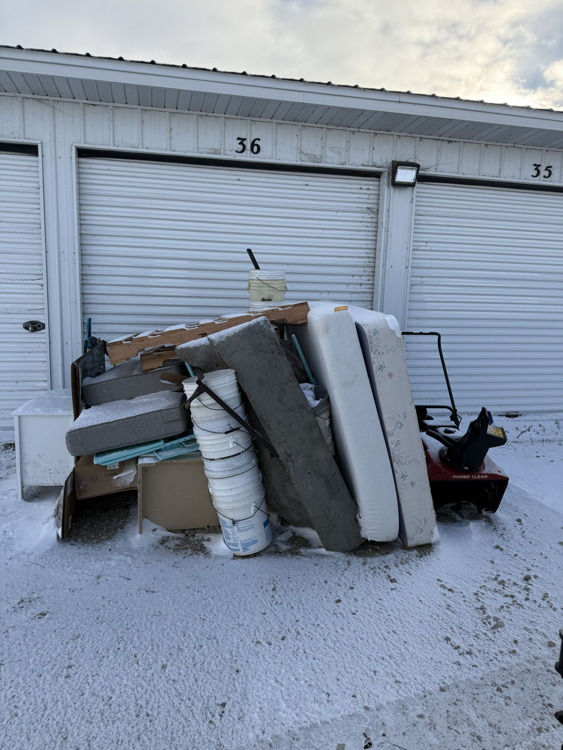 A pile of discarded items, including a mattress, cushions, and a red snowblower, left in front of storage units in the snow.
