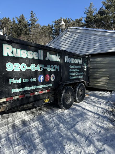 A black Russell Junk Removal trailer parked in the snow beside a white-sided building.
