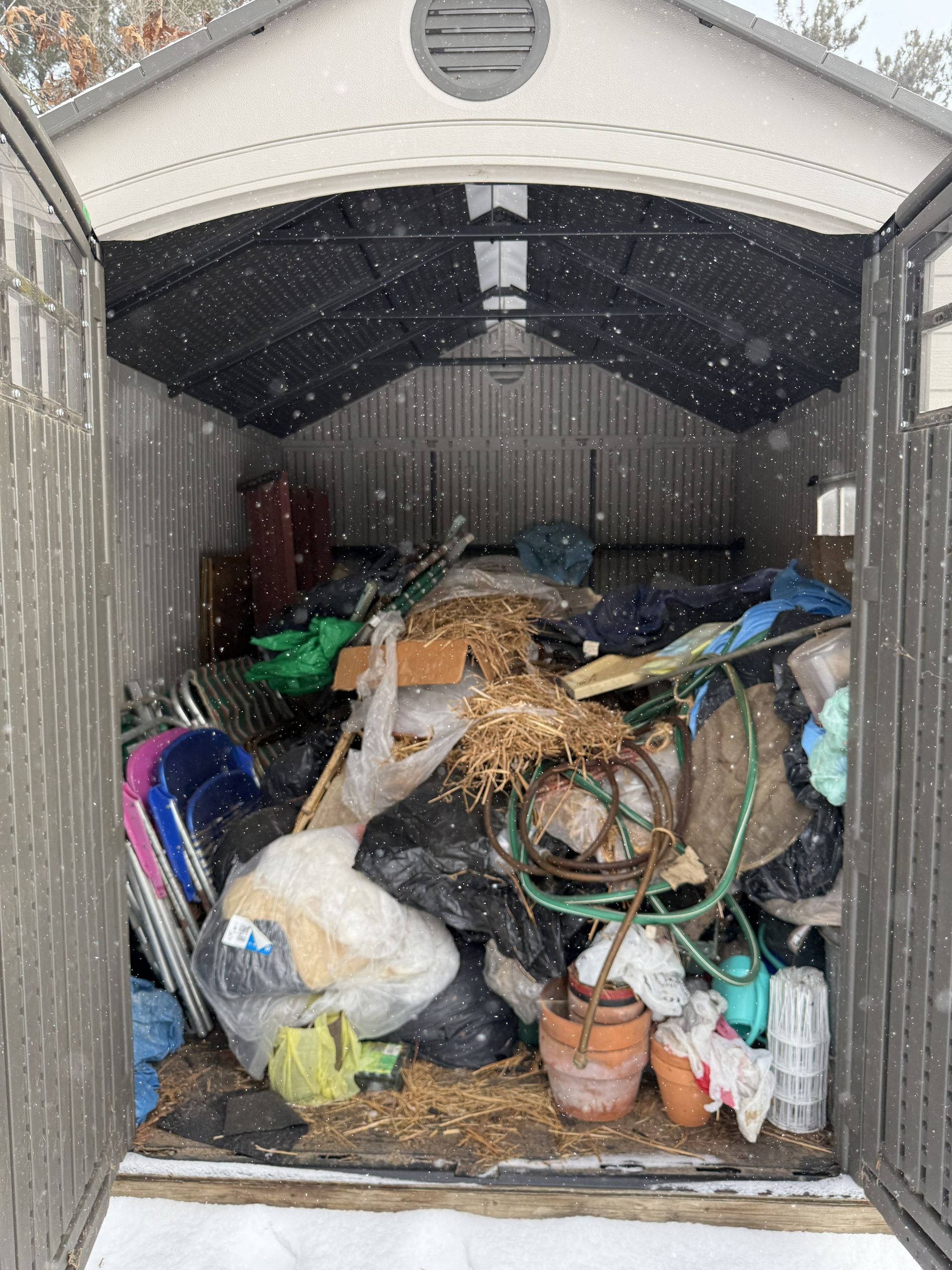 An open storage shed filled with miscellaneous household items, garden tools, and containers, viewed during a light snowfall.