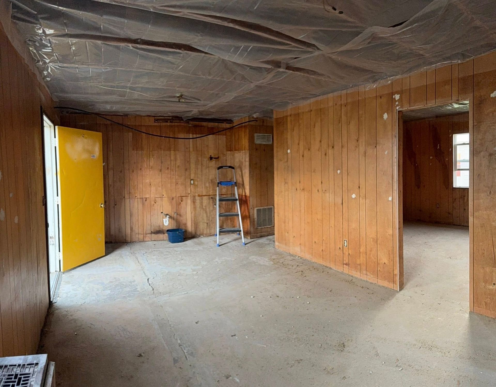 An interior room under construction with wood-paneled walls, a yellow door, a stepladder, and plastic-covered ceiling.