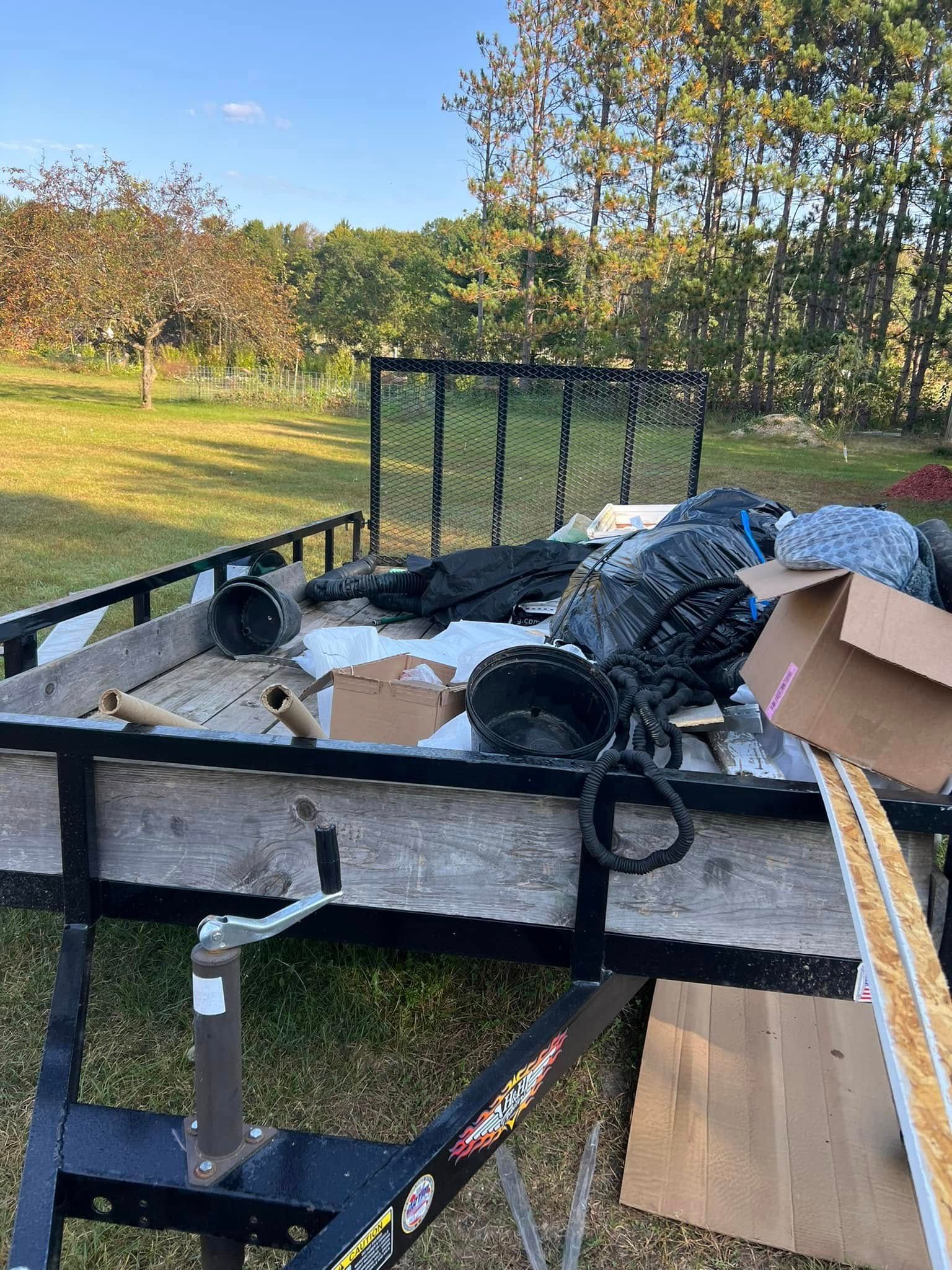A utility trailer sits outdoors in a field, loaded with cardboard boxes, trash bags, and various debris.