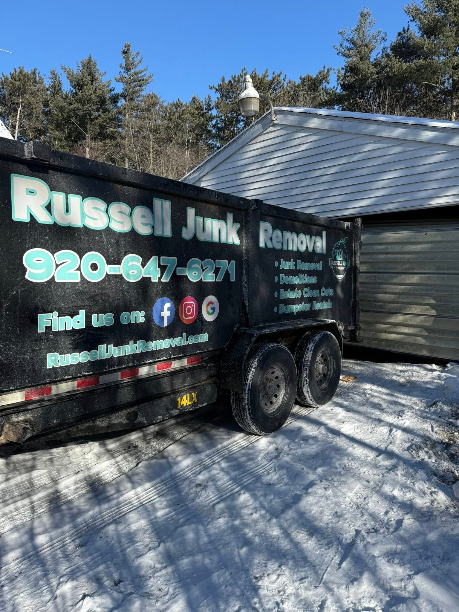 A black Russell Junk Removal trailer parked in the snow beside a white-sided building.