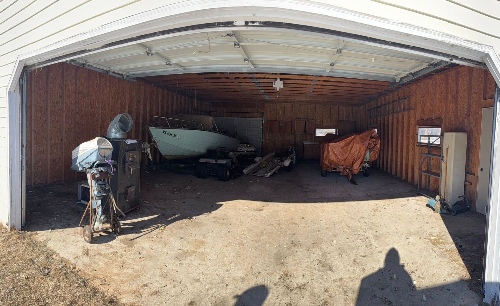 An open-faced garage containing a small boat, an outboard motor on a stand, and an object covered by an orange tarp.