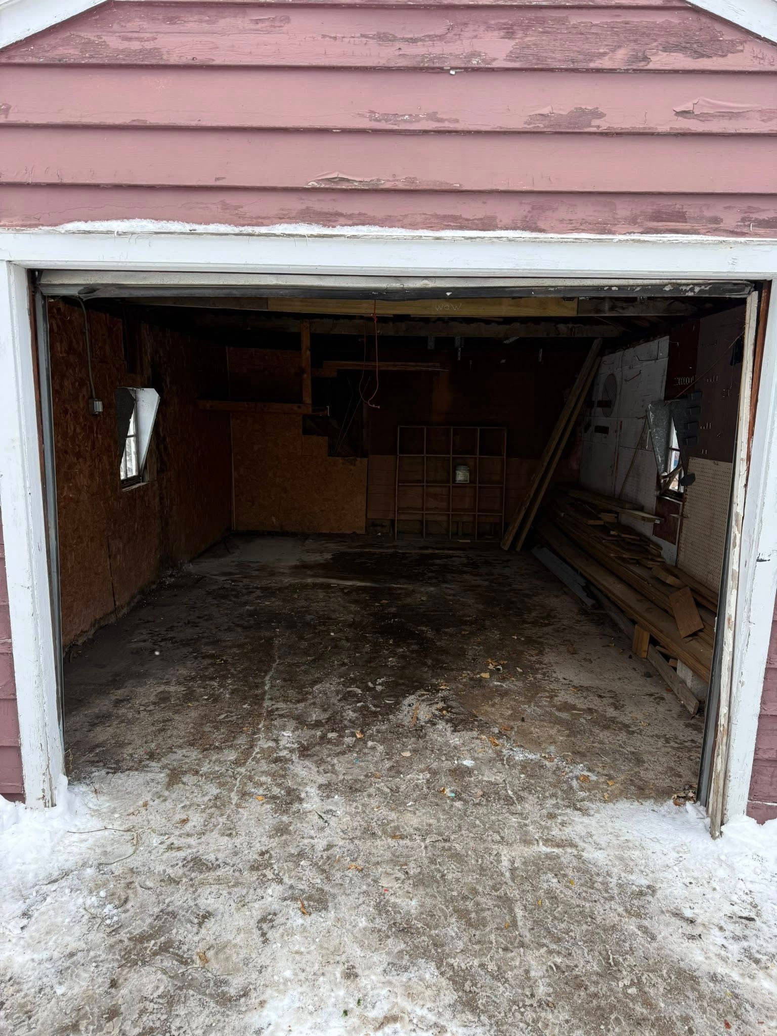An open, weathered garage with pink horizontal siding, dark interior walls, and snow on the ground at the entrance.