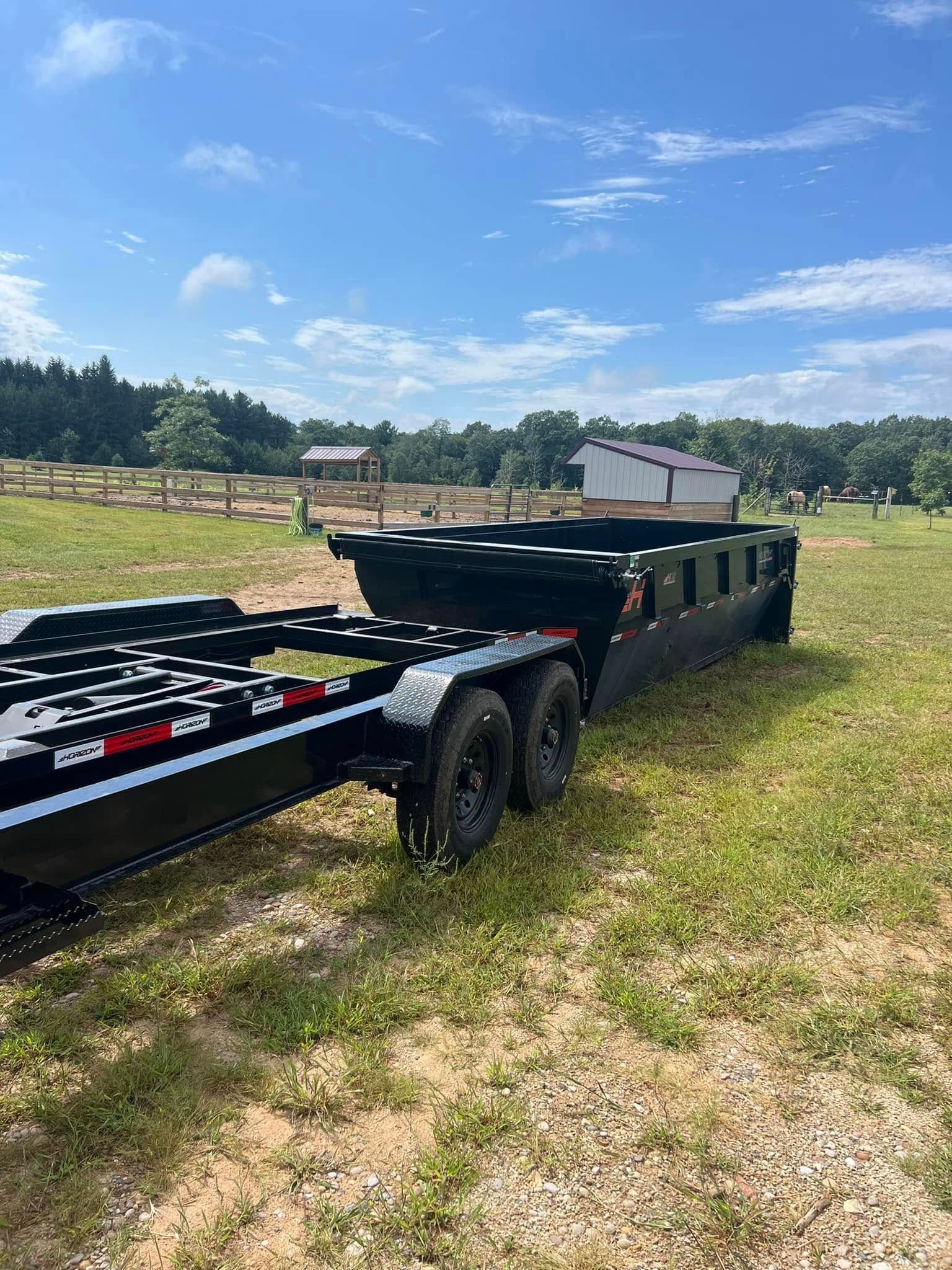 A black dump trailer sits on a grassy field under a sunny blue sky.